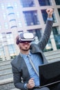 Happy young businessman using VR goggles, sitting in front of an office building. Celebrating success with fist high in the air Royalty Free Stock Photo