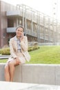 Happy young businesswoman using mobile phone while sitting on wall against office building Royalty Free Stock Photo