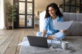 Happy young Cauasian lady working or studying on laptop computer, holding documents, looking at pc screen indoors