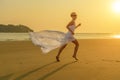 A happy young caucasian blonde woman in sunglasses and a fluttering white dress runs down the beach at sunset. Concept of Royalty Free Stock Photo