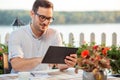 Young freelancer drinking coffee in riverside cafe and browsing internet on a tablet