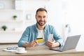 Happy young Caucasian man shopping online, using smartphone, laptop and credit card at home office Royalty Free Stock Photo