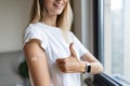 Happy young caucasian woman with blonde hair in white t-shirt showing patch on arm, feeling good after anti coronavirus