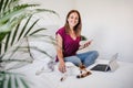 Happy young caucasian woman at home looking into camera working on laptop and mobile phone while cute jack russell dog resting on Royalty Free Stock Photo