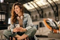 Happy young caucasian woman with phone and headphones is sitting at railway station.