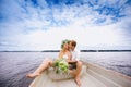 Happy young couple with a bouquet and a wreath hug sitting in a boat on the lake and sky background