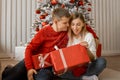 Happy young couple with gift boxes sitting near Christmas tree. Young man embrace his woman and woman slightly shows her tongue Royalty Free Stock Photo