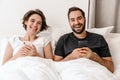 Happy young couple laughing and using mobile phones while lying in bed Royalty Free Stock Photo