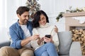 Happy young couple man and woman sitting on sofa together, hugging and watching video online using app on phone, joyful Royalty Free Stock Photo