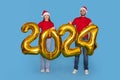 Happy Young Couple In Santa Hats Holding Golden 2024 Balloons In Hands