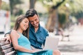 Happy young couple using laptop computer sitting on a bench in city outdoor - Two lovers having fun spending time together Royalty Free Stock Photo