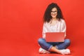 Happy young curly beautiful woman sitting on the floor with crossed legs and using laptop on red background Royalty Free Stock Photo