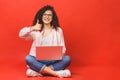 Happy young curly beautiful woman sitting on the floor with crossed legs and using laptop on red background. Thumbs up Royalty Free Stock Photo