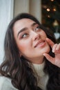 Happy young dark-haired curly woman in a light knitted sweater prepares gifts near the Christmas tree. Stylish model makes online