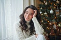 Happy young dark-haired curly woman in a light knitted sweater prepares gifts near the Christmas tree. Stylish model makes online