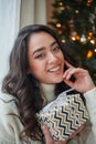 Happy young dark-haired curly woman in a light knitted sweater prepares gifts near the Christmas tree. Stylish model makes online