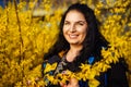 Happy young dark-haired plus size woman smiling happily in spring among yellow flowering forsythia bushes Royalty Free Stock Photo