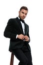 Happy young elegant man sitting on wooden chair fixing black tuxedo Royalty Free Stock Photo