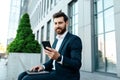 Happy young european businessman with beard in suit typing on phone and laptop, has chat with client