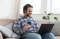 Happy young european male shopaholic with stubble in shirt shows credit card and looks at laptop sits on sofa