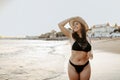 Happy young european woman in swimswear and straw hat standing on beach while walking along coastline at sunset Royalty Free Stock Photo