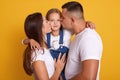 Happy young family kissing ther little charming daughter. Cute child girl sitting close to parents and looks at camera. Mother,