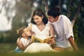 Happy young family reading a book in nature under a tree. Dad and little son fool around and show language to each other