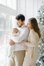 A happy young family with a small child are relaxing by the big window and the Christmas tree at home. Mother, daughter in a Royalty Free Stock Photo