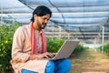 Happy young farmer busy working on laptop while sitting at greenhouse - concept of modern farming, technology and