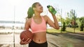 Happy young female basketball player smiling aside while drinking water from bottle, holding basketball, standing Royalty Free Stock Photo