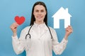 Happy young female doctor in professional medical white uniform and stethoscope holds small house and red heart, looking at camera Royalty Free Stock Photo