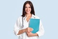 Happy young female family doctor holds blue folder with documents, has friendly expression, going to have look at patient, gives s