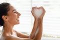 Happy young female sitting in bubble bath, making foamy heart with her hands at home