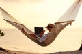 Happy young female wearing straw hat waving while having video call on laptop, relaxing in the hammock on tropical beach Royalty Free Stock Photo