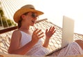 Happy young female wearing straw hat waving while having video call on laptop, relaxing in the hammock on tropical beach