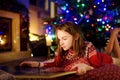 Happy young girl reading a story book by a fireplace in a cozy dark living room on Christmas eve. Celebrating Xmas at home