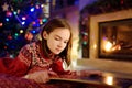 Happy young girl reading a story book by a fireplace in a cozy dark living room on Christmas eve. Celebrating Xmas at home
