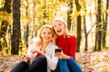 Happy young girls holding mobile phone and sitting on fallen leaves outdoor. Young women in Autumn. Students friends on Royalty Free Stock Photo