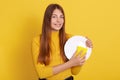 Happy young good looking woman washing dishes, posing  over yellow background, showing white plate and sponge, looking at Royalty Free Stock Photo