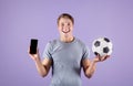 Happy young guy holding soccer ball and smartphone with blank screen on lilac studio background, mockup for design
