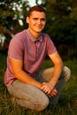 Happy young guy. Portrait of smiling young brunette man in purple polo shirt seating outdoor on nature background at Royalty Free Stock Photo