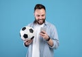 Happy young guy with soccer ball using smartphone, winning sports bet, rooting for his favorite team on blue background Royalty Free Stock Photo