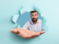 Happy young guy stretching open palm through hole in torn blue paper, offering helping hand, reaching out for support Royalty Free Stock Photo