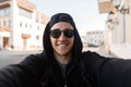 Happy young hipster man in stylish sunglasses in a black hoodie in a baseball cap with a cute smile makes a selfie Royalty Free Stock Photo