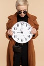 Happy young hipster woman with blonde short hair wearing a coat and sunglasses holding a clock over beige background. Royalty Free Stock Photo