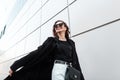 Happy young hipster woman in fashionable long black coat in white jeans in stylish sunglasses with a leather bag travels