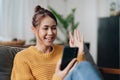 Happy young hispanic asian teen girl sit on sofa at home holding phone looking at screen waving hand video calling Royalty Free Stock Photo