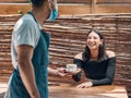 Happy young hispanic woman receiving a cup of coffee from a waiter wearing a face mask in a local cafe. Mixed race woman Royalty Free Stock Photo