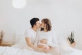 Happy young husband kissing his smiling pregnant wife and hugging belly bump on white bed. Stylish pregnant couple in white Royalty Free Stock Photo