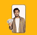 Happy young indian guy with open mouth show many dollar in hand on empty screen of big phone